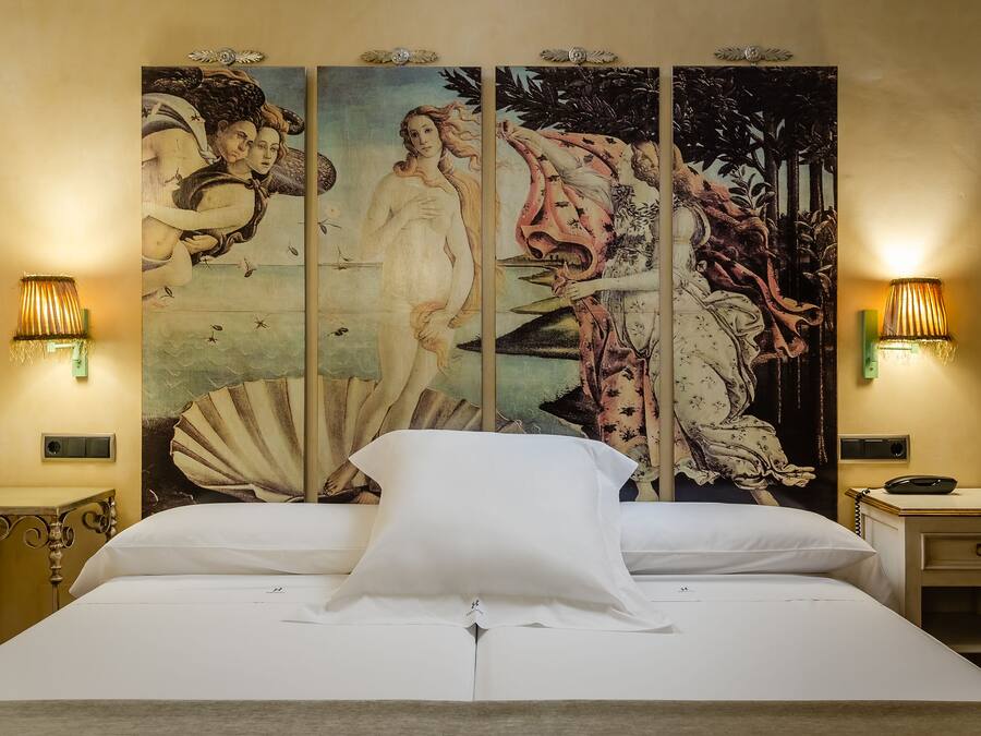 A bed with white linens, flanked by two nightstands with lamps, and a large, ornate tapestry on the wall behind.