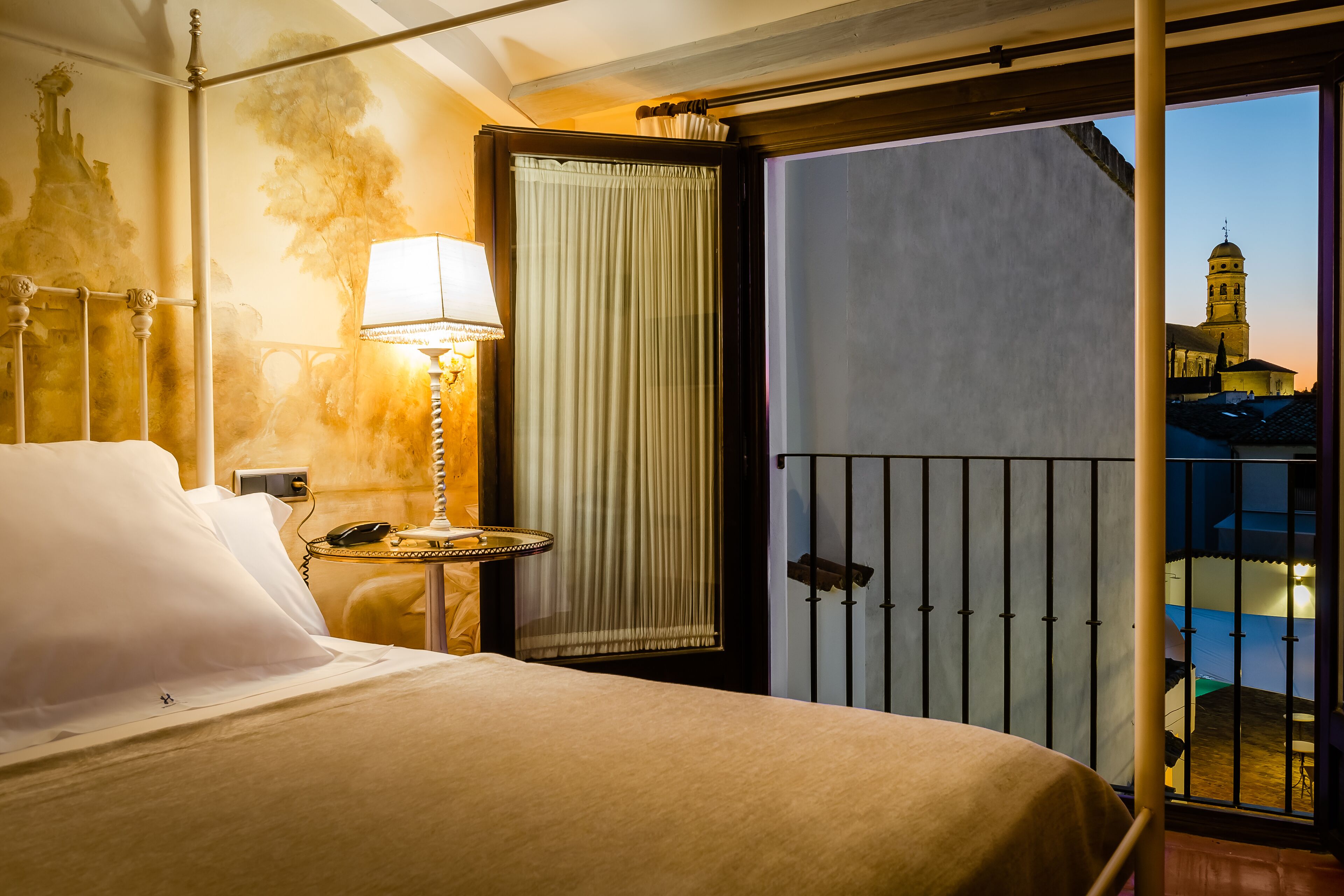 Habitación de hotel con cama, mesita de noche con lámpara, cortina y balcón con vistas a un edificio.