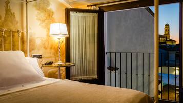 Habitación de hotel con cama, mesita de noche con lámpara, cortina y balcón con vistas a un edificio.