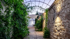 A narrow stone pathway flanked by lush greenery and a stone wall, leading to a covered area with tables and chairs. - Hotel  YIT La Casona del Arco (Baeza)