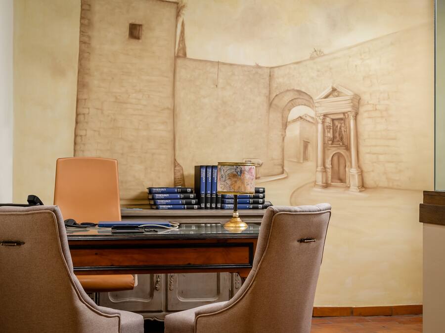 Una sala de estudio con un escritorio, dos sillas, una estantería llena de libros y un mural de una escena arquitectónica.
