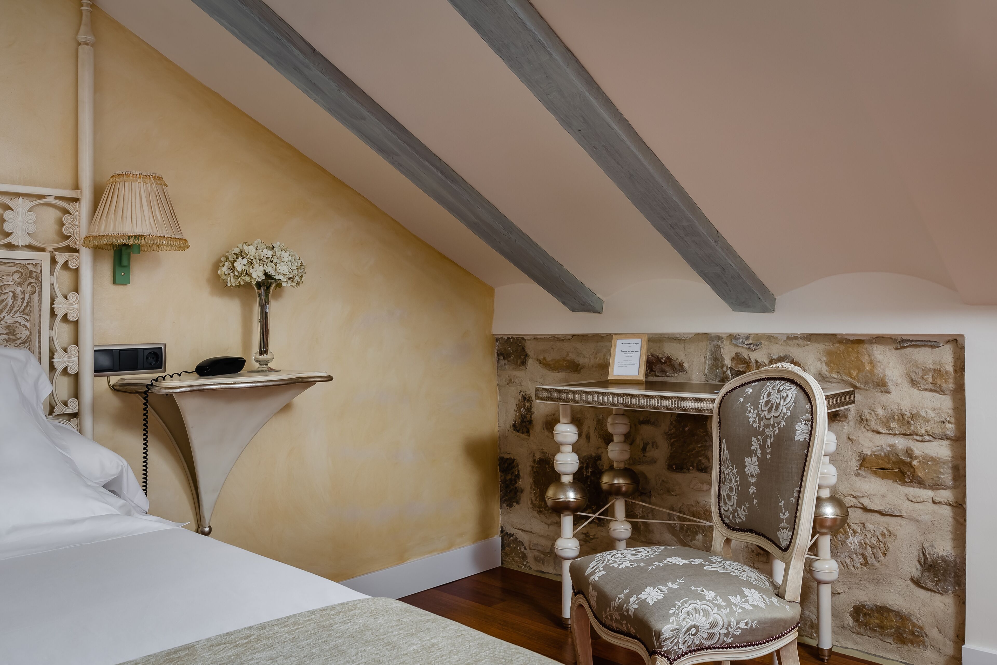 Ein Schlafzimmer mit einem Bett, einem Nachttisch mit Lampe, einem Sessel und einer Steinwand.