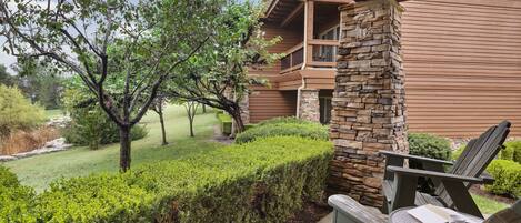 Un edificio de madera con un pilar de piedra, un balcón y un jardín bien cuidado con un banco y sillas.