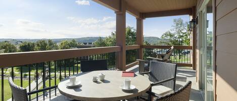 Un balcon agrémenté de meubles en osier, une table avec des tasses et un livre, et une vue panoramique sur des collines et des arbres.