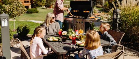 A family dining outdoors with a grill, a table, and chairs.