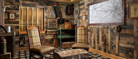 A rustic room with wooden walls, a large map, a green table, two plaid chairs, and a trunk with a lock.