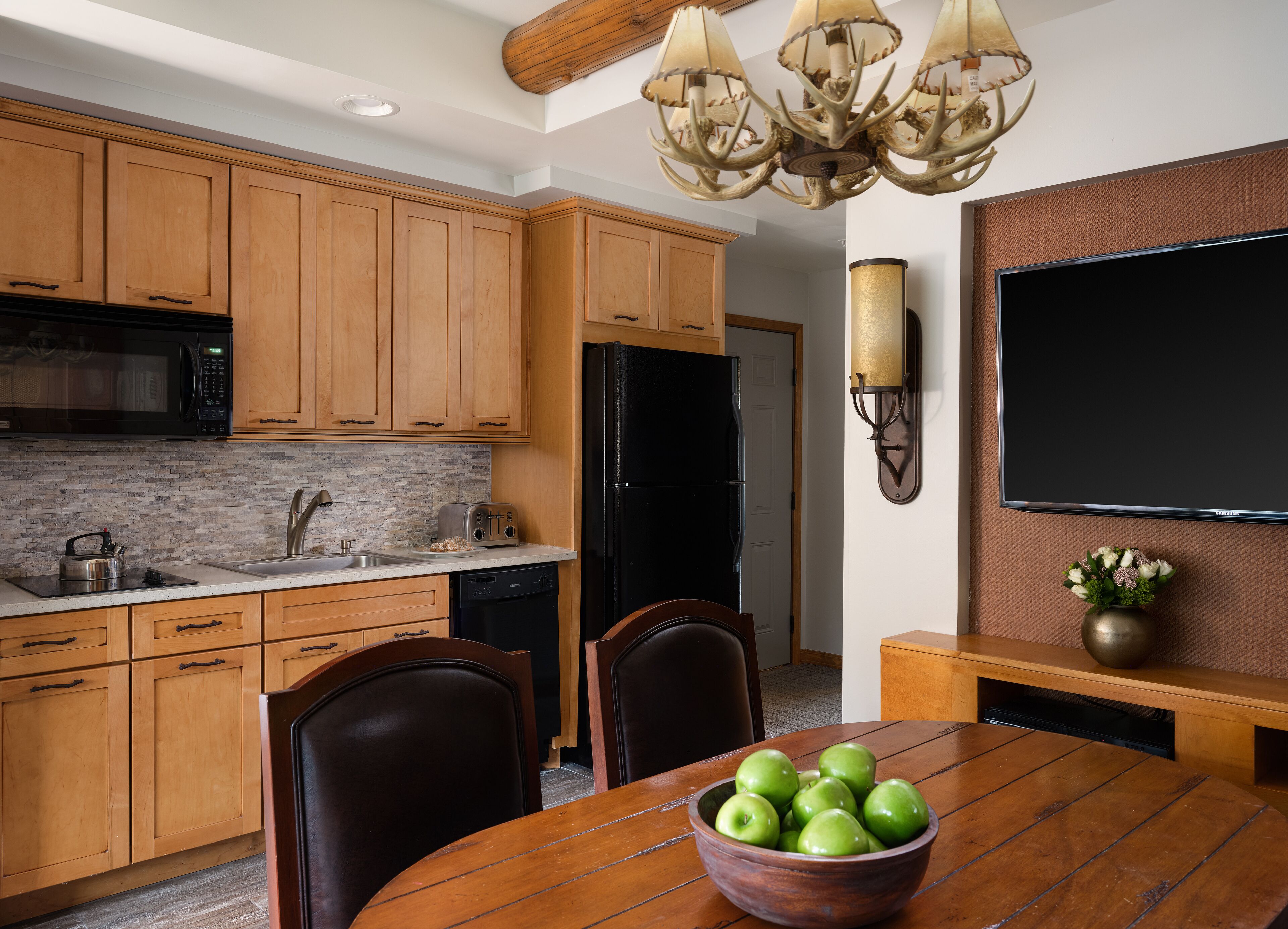 A modern kitchen with wooden cabinets, a black refrigerator, a microwave, a toaster, and a dining table with a bowl of green apples.