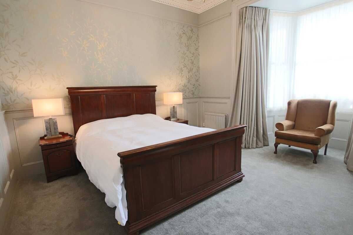 A neatly arranged bedroom with a large wooden bed, two bedside tables with lamps, a chair, and a window with curtains.
