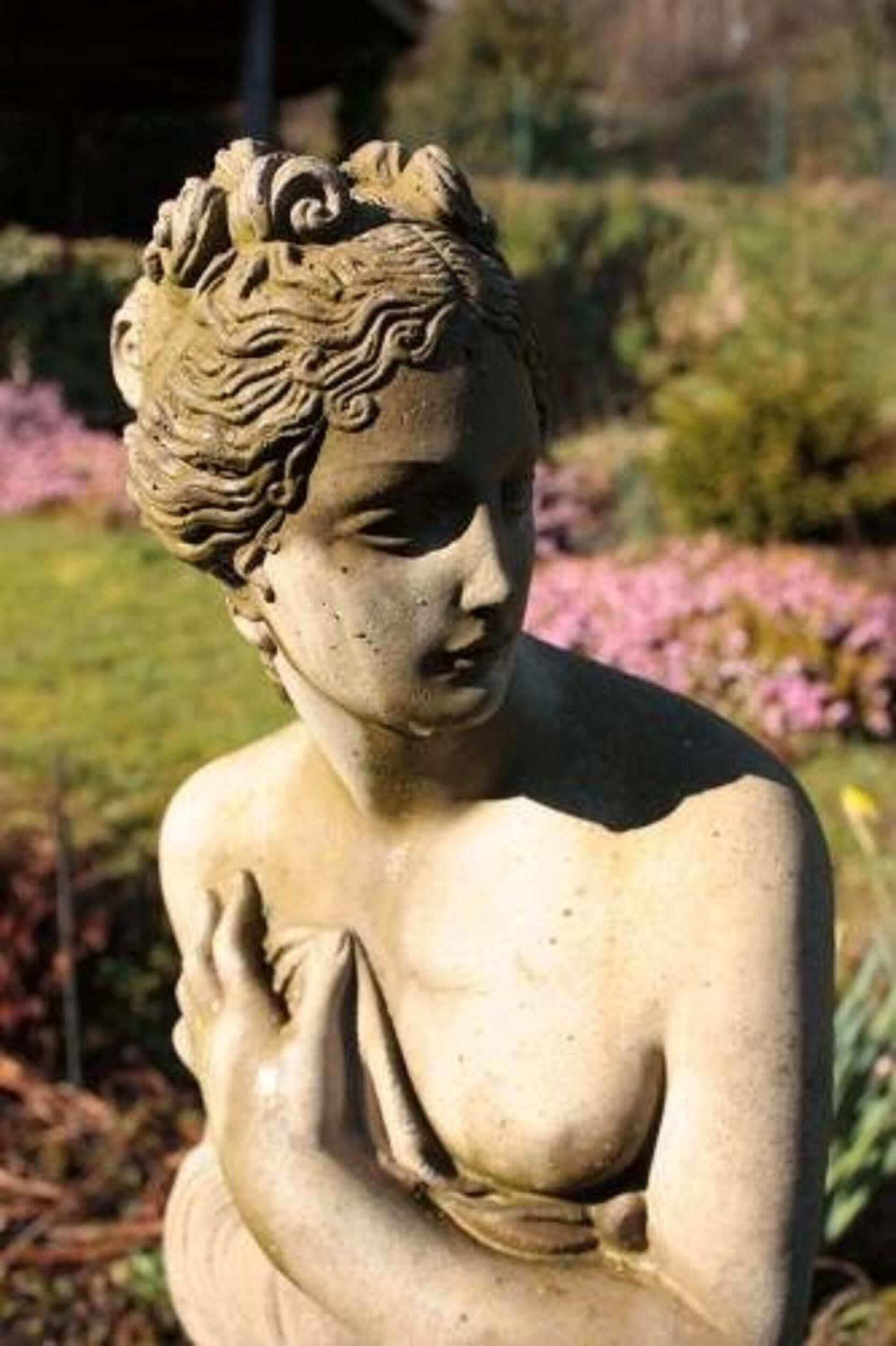 A statue of a woman with curly hair, set in a garden with flowers and grass.