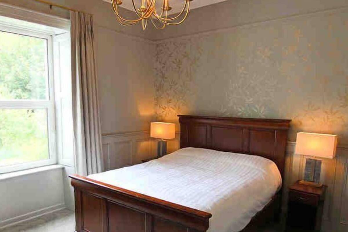 A bedroom with a large bed, two bedside tables with lamps, a chandelier, and a window with curtains.