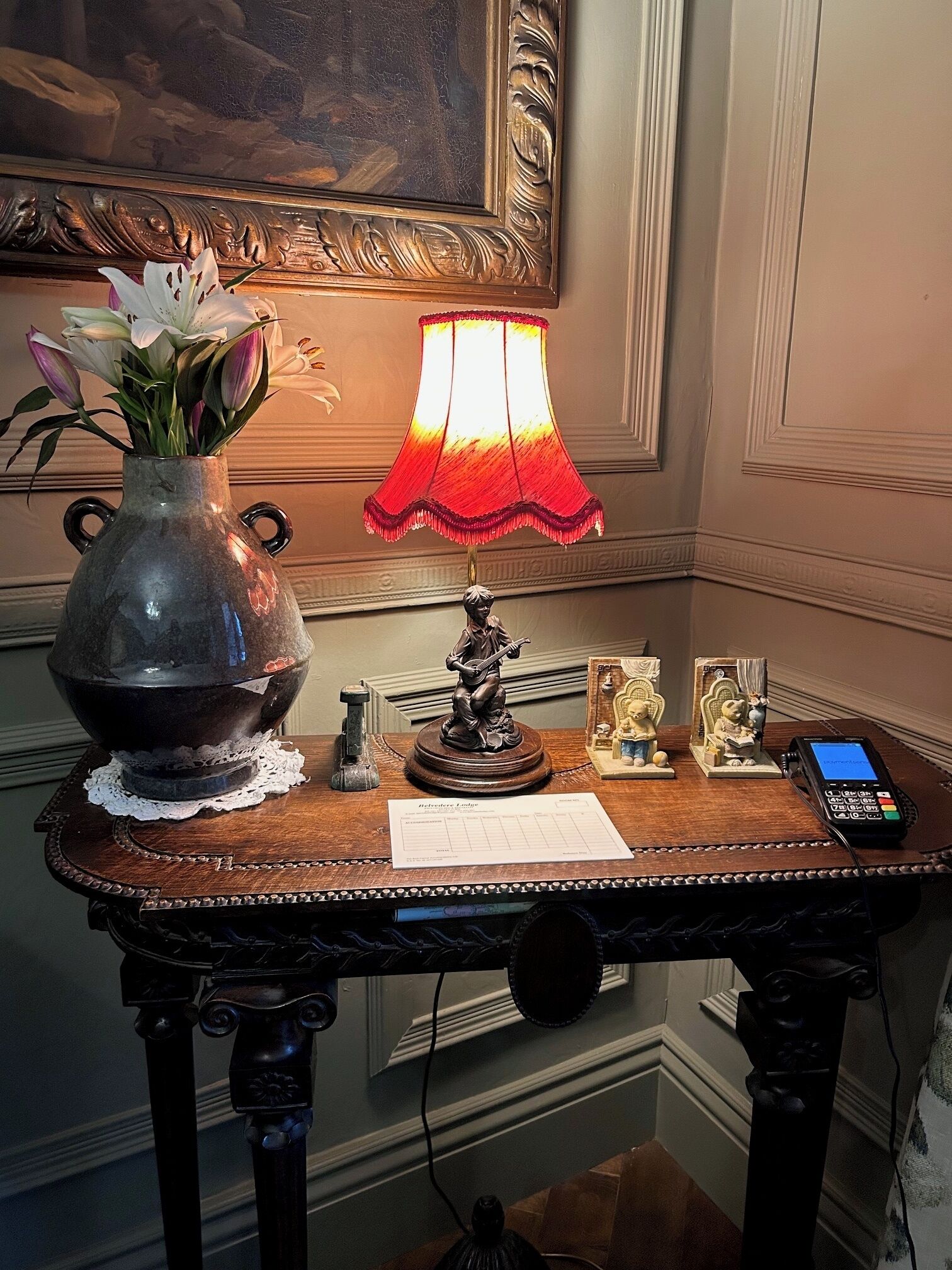 A wooden table with a vase of flowers, a lamp, a statue, and a payment terminal.