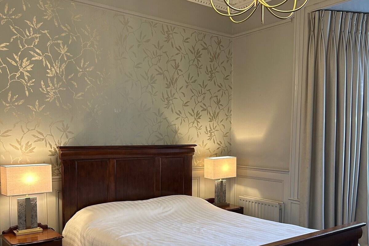 A bedroom with a large bed, two bedside tables with lamps, a radiator, and a window with curtains.