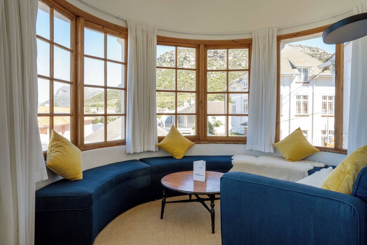 Eine Sitzecke mit gebogenem, blauen Sofa, einem runden Holztisch und großen Fenstern mit Blick auf Häuser und Hügel.