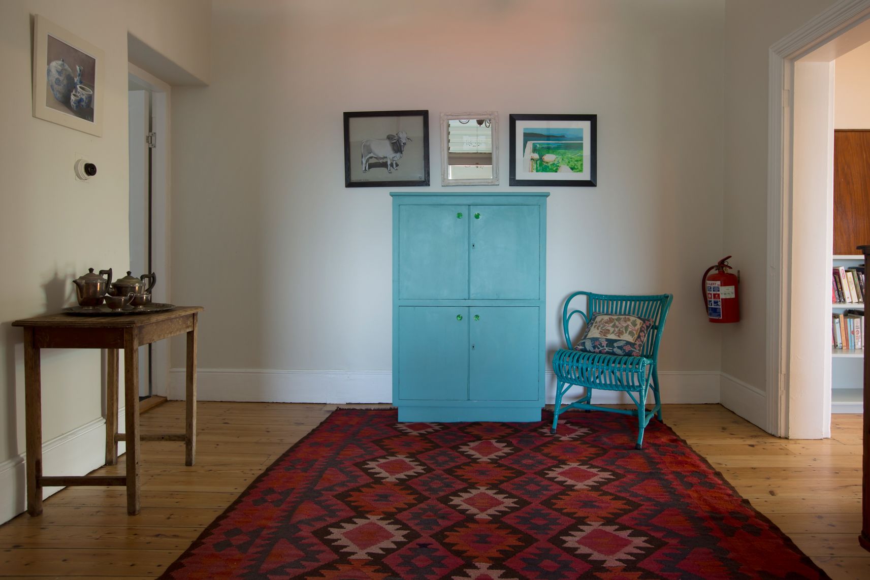 Una habitació amb sòl de fusta, una catifa amb dibuixos, un armari color turquesa, una cadira de vimbles i una biblioteca.