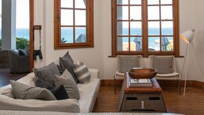 A room with a white sofa, two wicker chairs, a wooden coffee table, and a view of the sea through large windows.