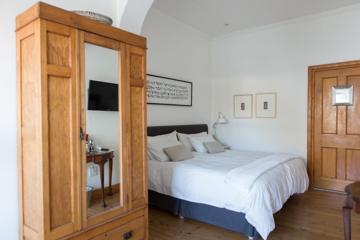 Un dormitorio con un armario de madera, una cama con ropa de cama blanca, una mesita de noche, una lámpara y un televisor fijado a la pared.