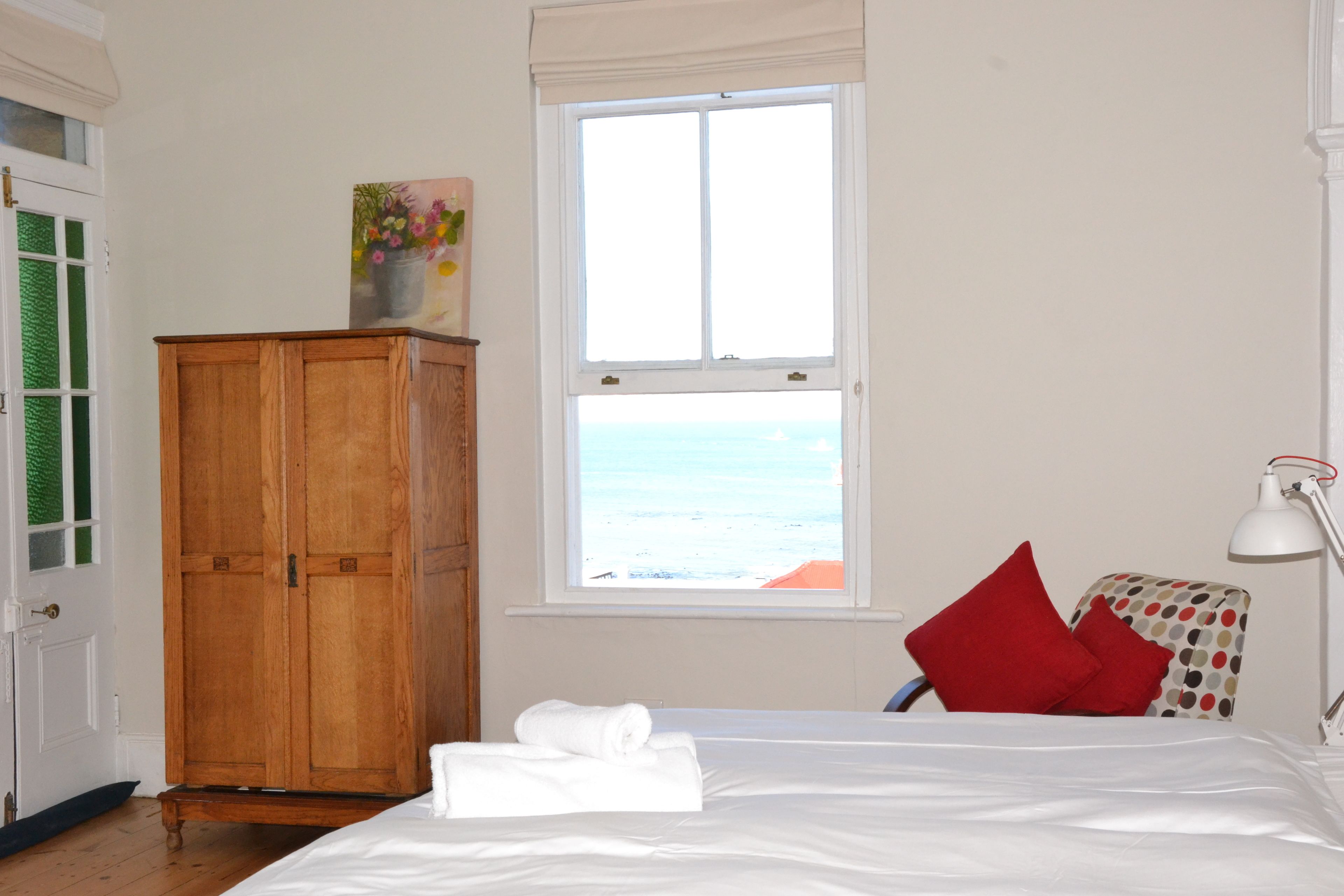 Een slaapkamer met een bed, een houten garderobe, een raam met uitzicht op zee en een bureau met een lamp.