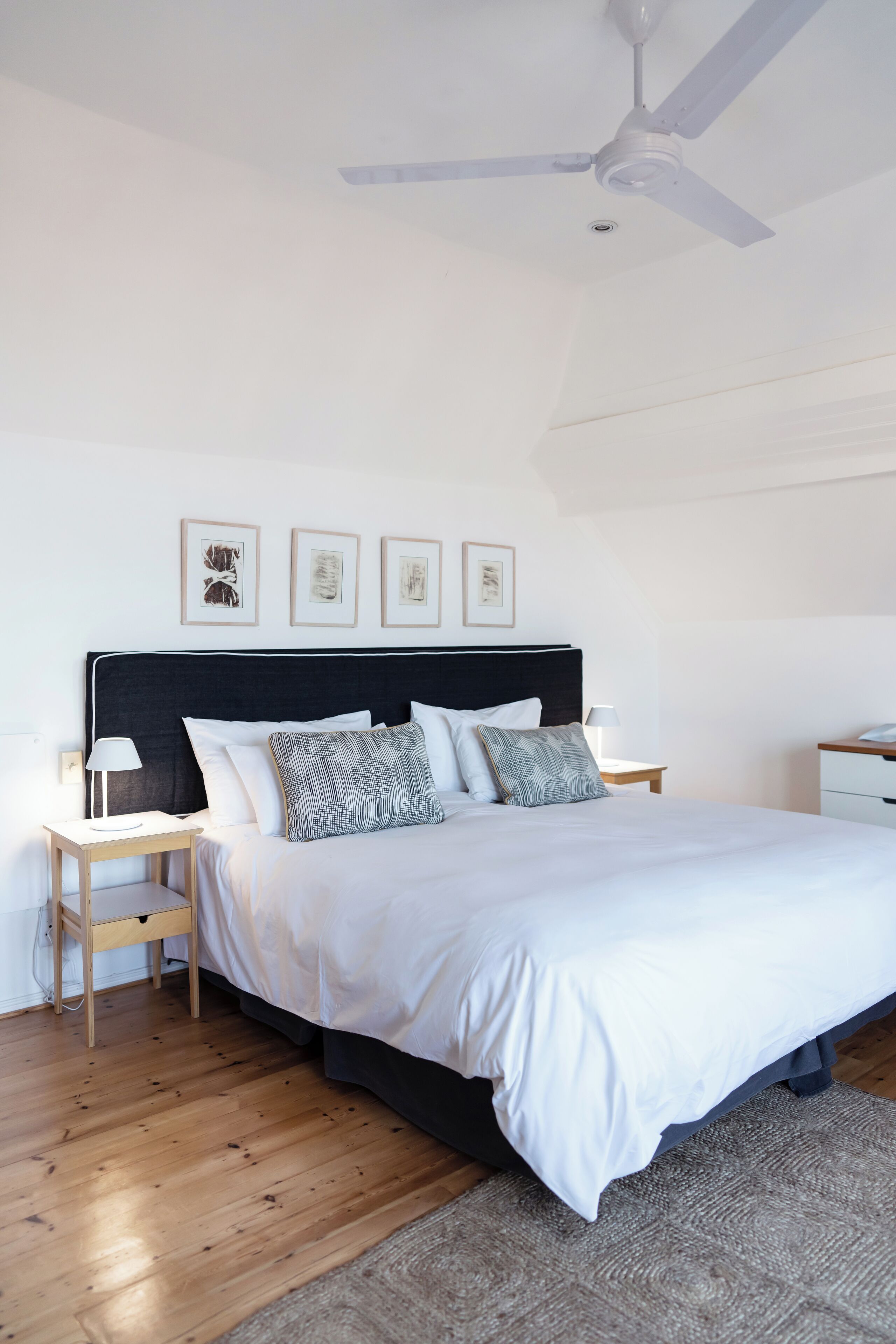 A bedroom with a bed, wooden floor, ceiling fan, bedside tables, and framed pictures on the wall.