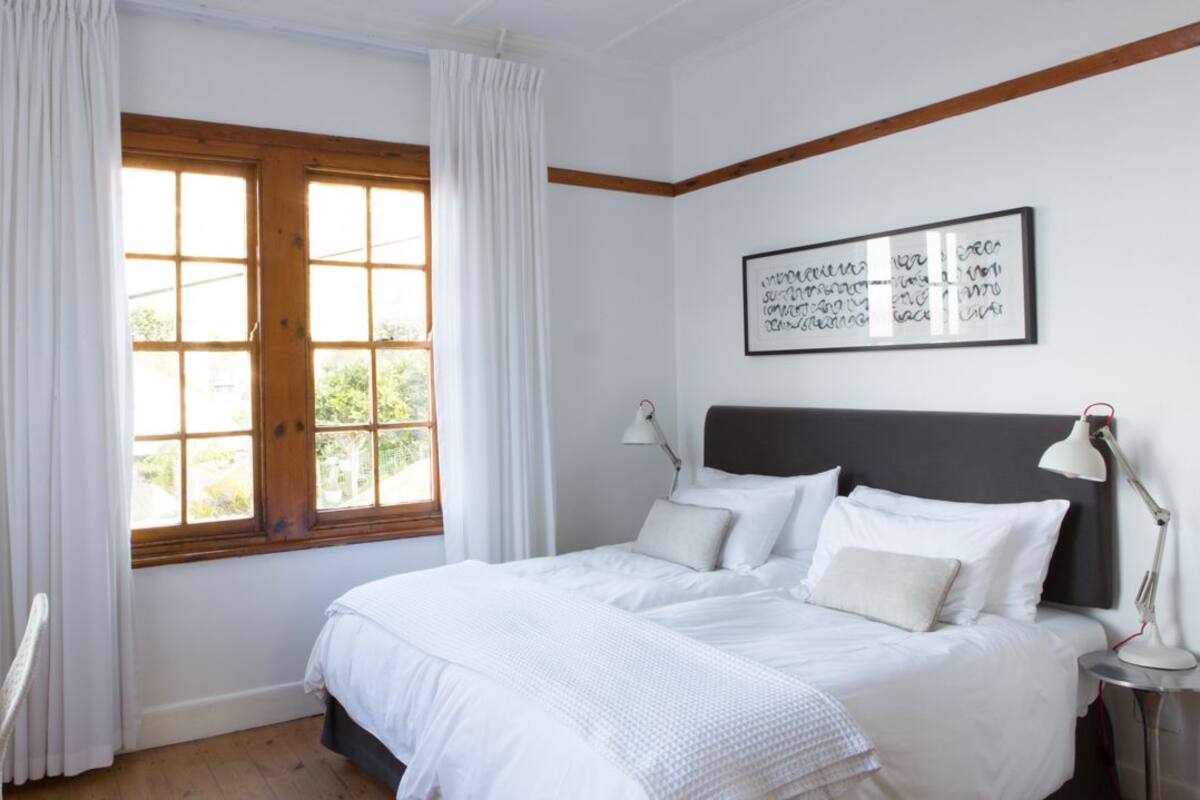 Ein Schlafzimmer mit großem Fenster, einem Bett mit weißer Bettwäsche, einem Nachttisch mit Lampe und einem gerahmten Kunstwerk an der Wand.