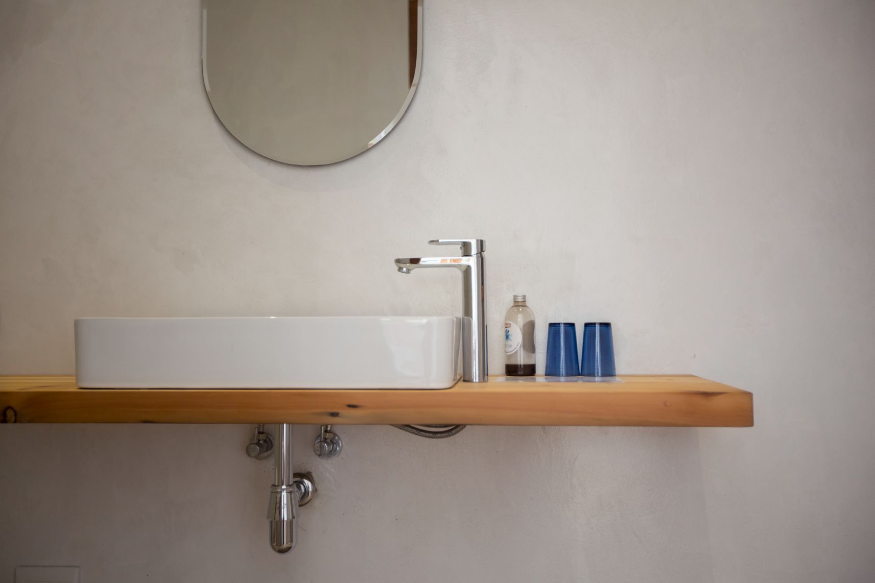 A bathroom with a white sink, a mirror, and a wooden shelf with two blue cups and a bottle.