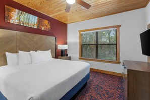 A hotel room with a large bed, a headboard, a window with blinds, a bedside table with a lamp, and a flat-screen TV. - WorldMark New Braunfels (New Braunfels)
