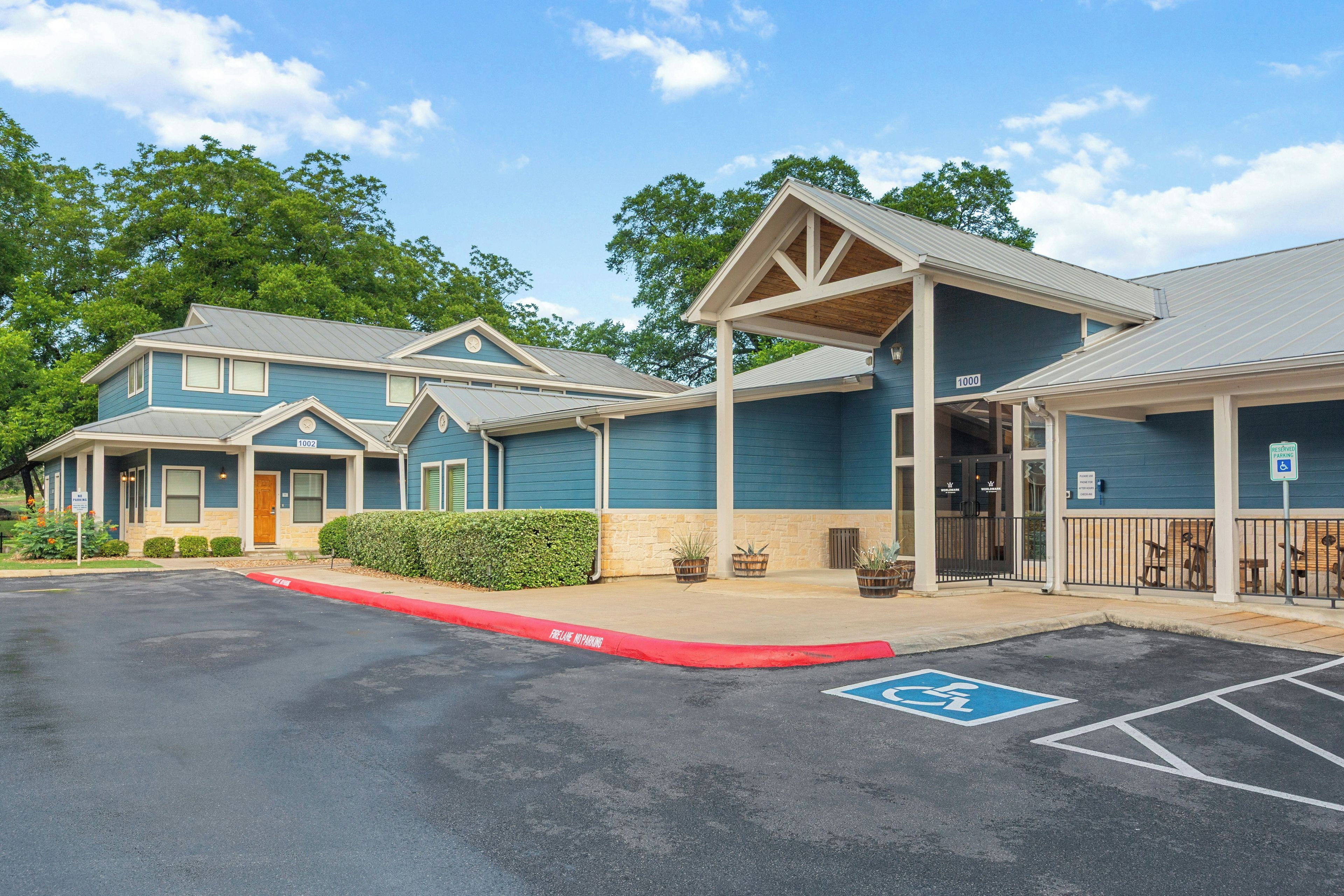 A row of blue buildings with numbered doors, a parking lot with a red curb, and a blue handicapped parking sign.
