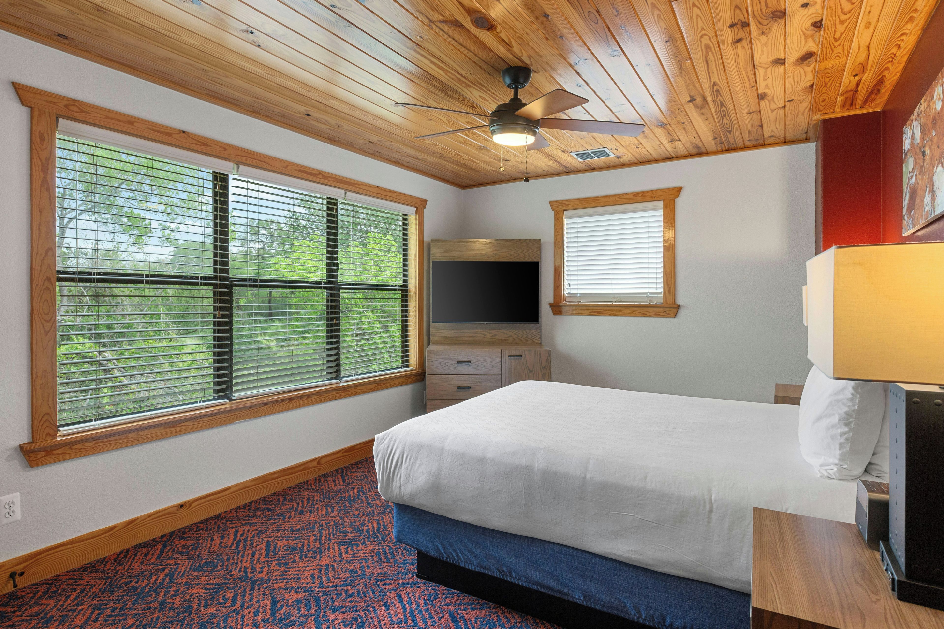 A bedroom with a bed, a ceiling fan, a window with blinds, a dresser, and a nightstand with a lamp.
