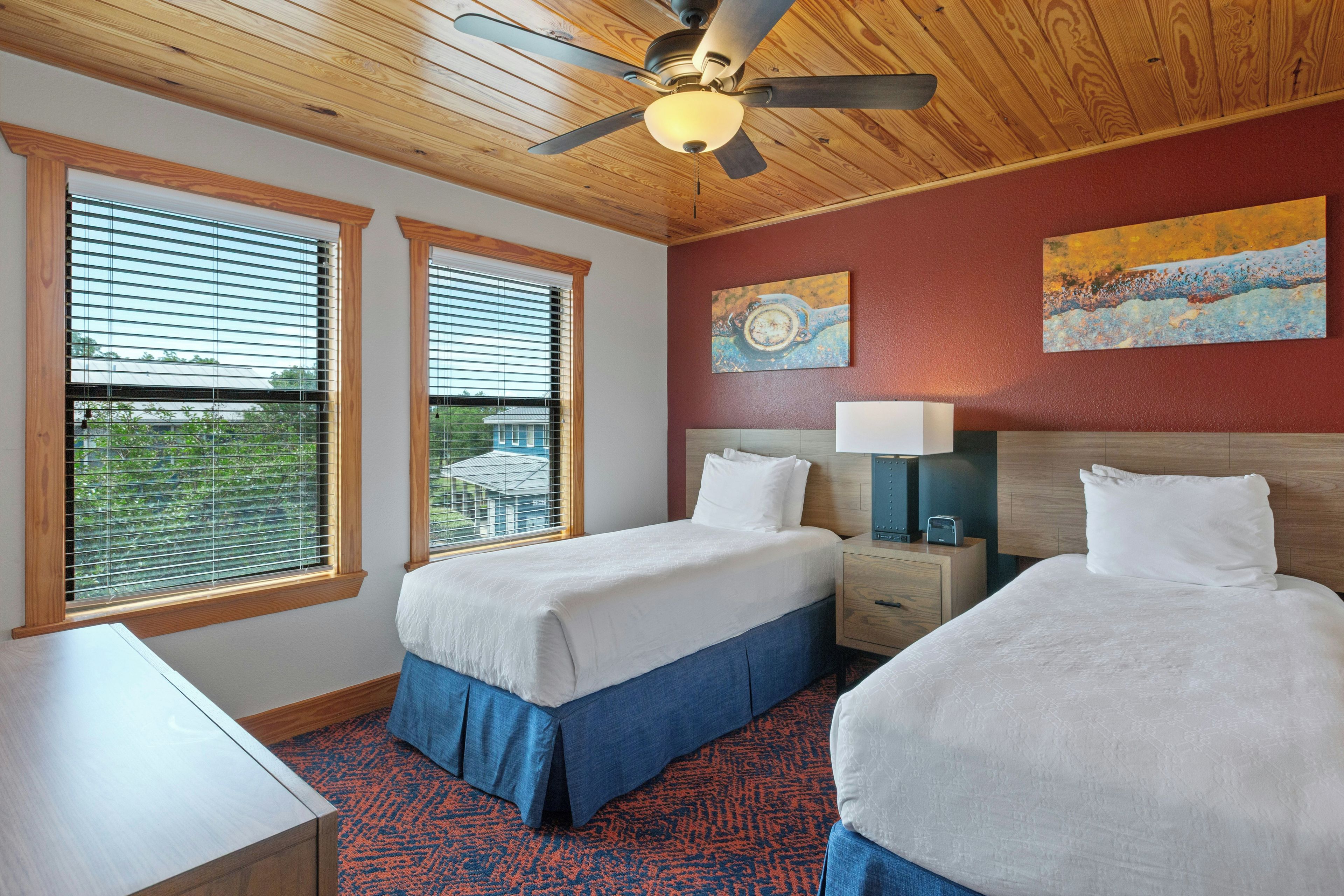 A hotel room with two beds, a ceiling fan, a window with blinds, and artwork on the wall.