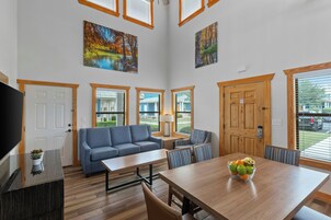 A spacious living area with a sofa, a dining table, and a TV. There are paintings on the walls and a view of the outdoors through the windows.