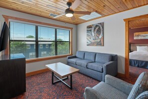 A room with a ceiling fan, a large window with blinds, a gray sofa, a small table, and a bed with a wooden headboard.