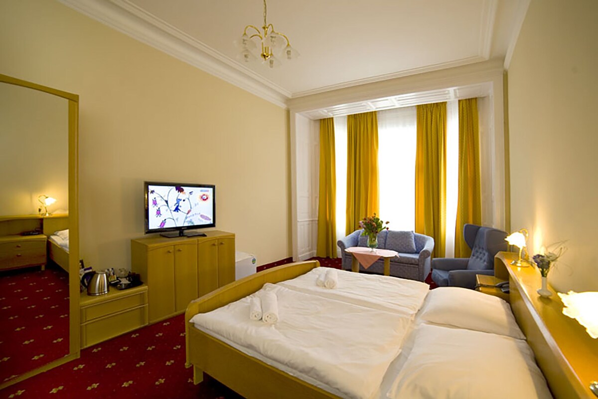 A hotel room with a bed, a television, a desk with a lamp, a chair, and a window with curtains.