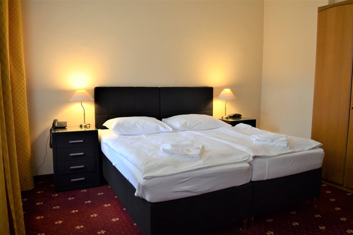 A neatly made double bed with white linens, two bedside tables with lamps, a telephone, and a small drawer unit.