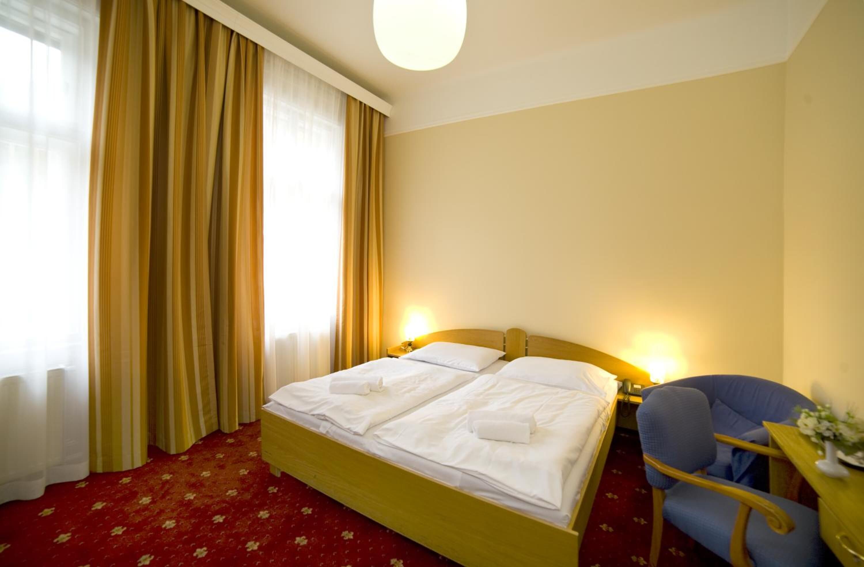 A hotel room with a double bed, two bedside lamps, a blue armchair, and a small table with a vase.