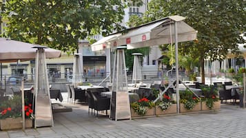 An outdoor cafe with tables and chairs, covered by umbrellas, and surrounded by plants and flowers.