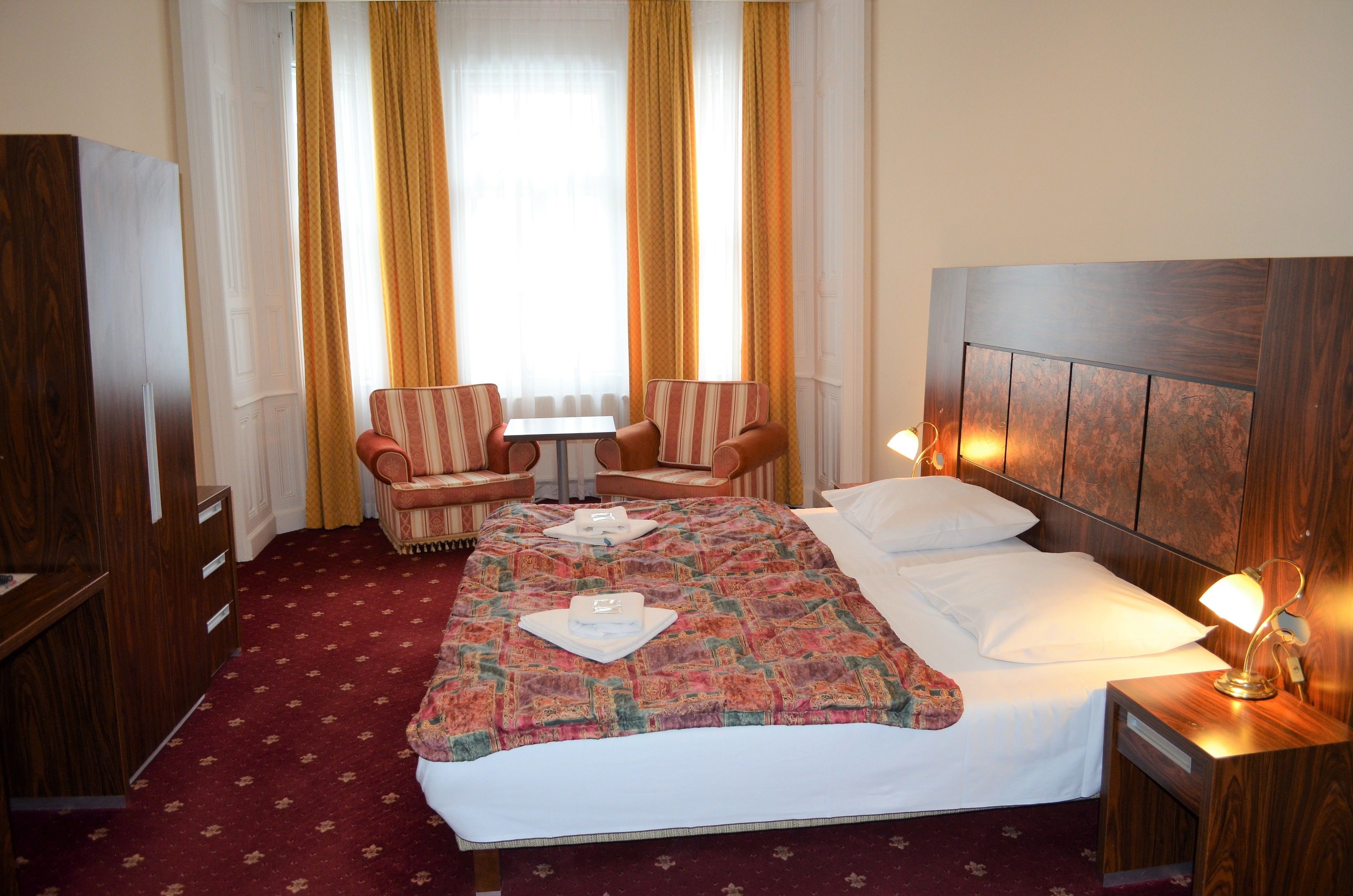 Een hotelkamer met een groot bed, twee fauteuils, een kleine tafel en een nachtkastje met een lamp.
