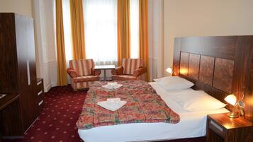 Een hotelkamer met een groot bed, twee fauteuils, een kleine tafel en een nachtkastje met een lamp.