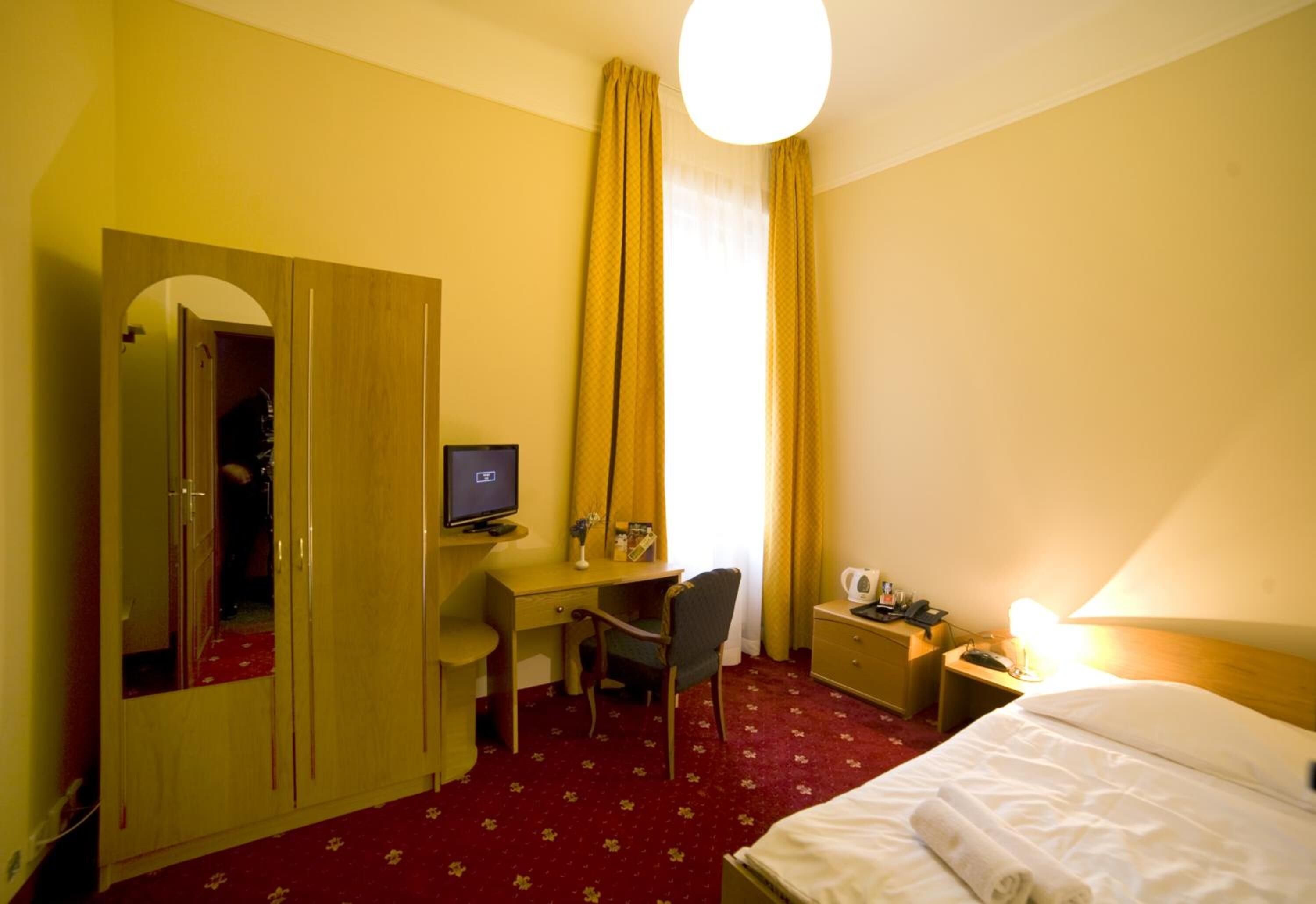 Habitación de hotel con cama, escritorio con computadora, silla, armario, mesita de noche con lámpara y ventana con cortinas.