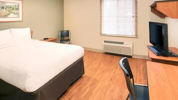 A hotel room with a bed, a desk with a computer, a chair, a TV, and a window with blinds.
