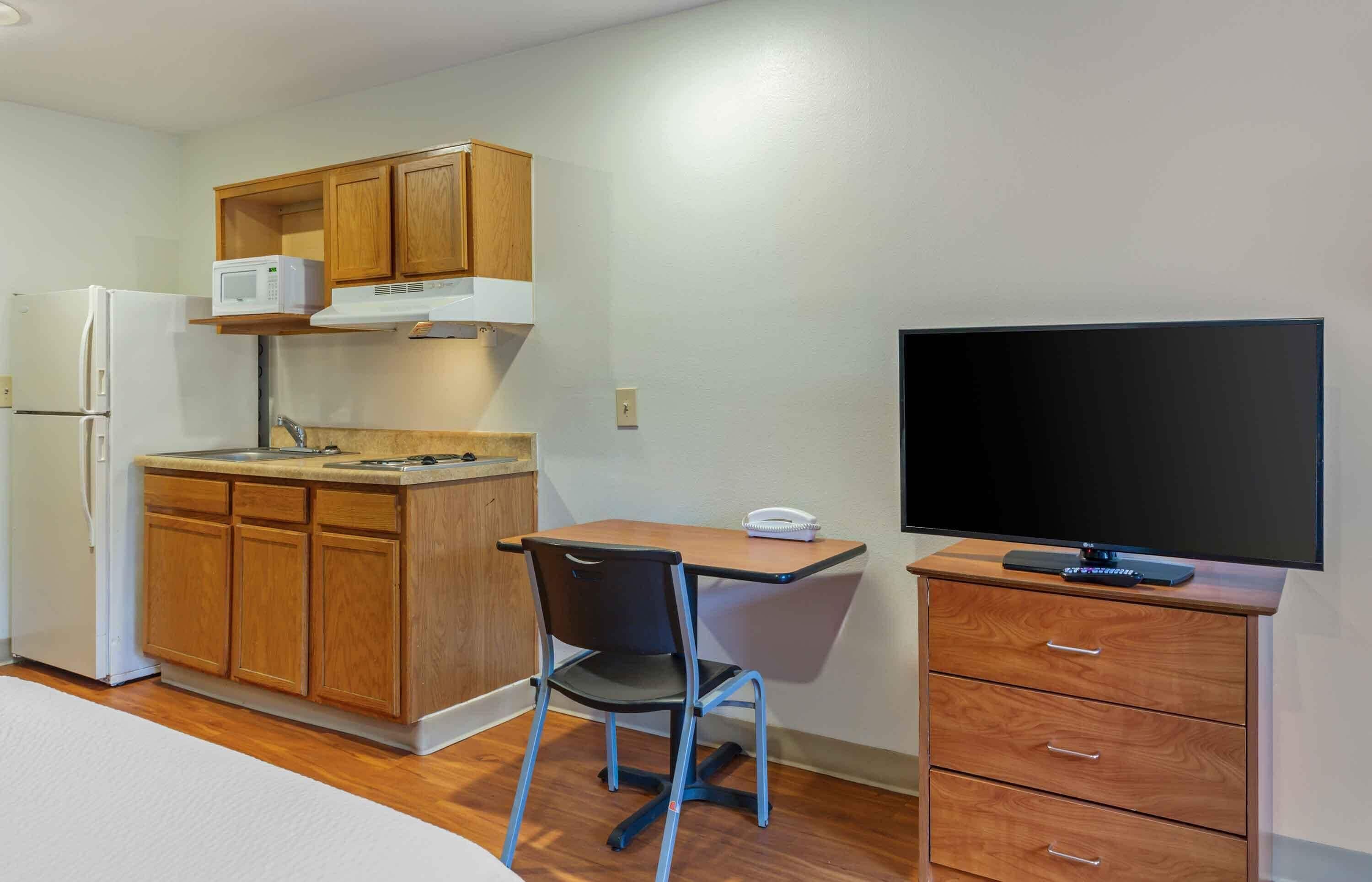 A compact kitchenette with a microwave, stove, and refrigerator. There is a small dining table with a chair, a television on a wooden cabinet, and a drawer unit.