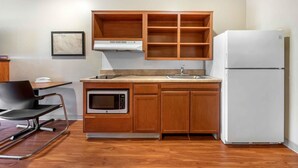 A compact kitchen with wooden cabinets, a built-in microwave, a white refrigerator, and a small dining area with a chair and table. - Extended Stay America Select Suites - Fayetteville - West (Fayetteville)