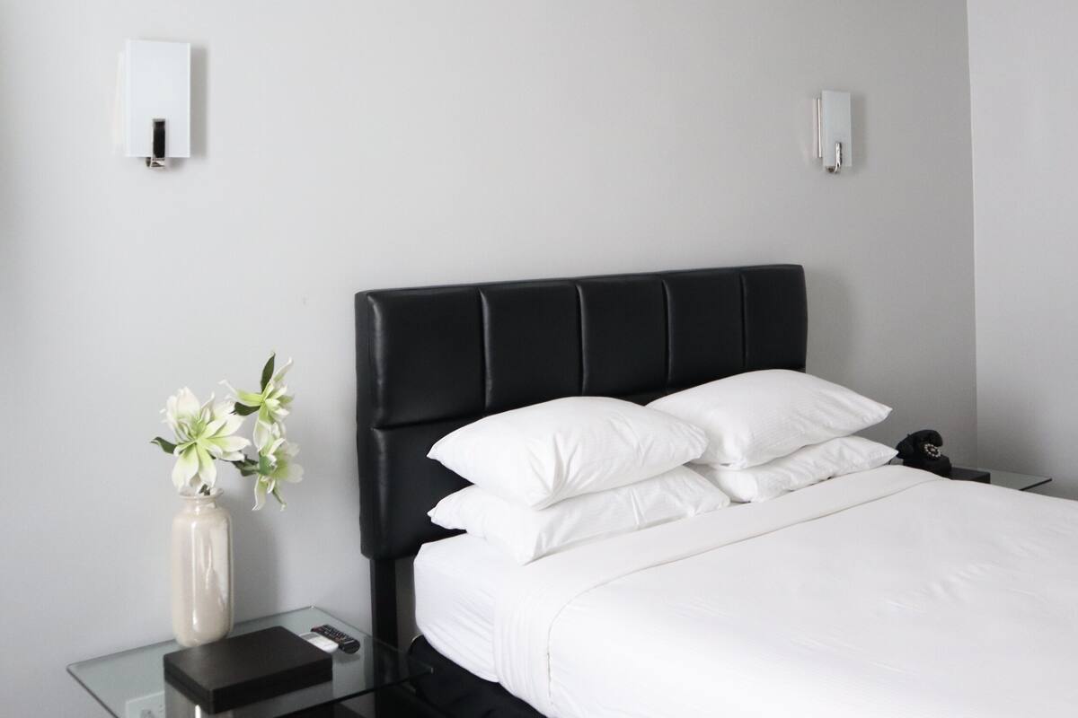 A hotel room with a black upholstered headboard, white bedding, a bedside table with a vase of flowers, and a bedside lamp.