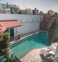 Una piscina con una estructura de techo rojo, un área de estar cubierta y un edificio blanco con un letrero que dice @TITUDE.