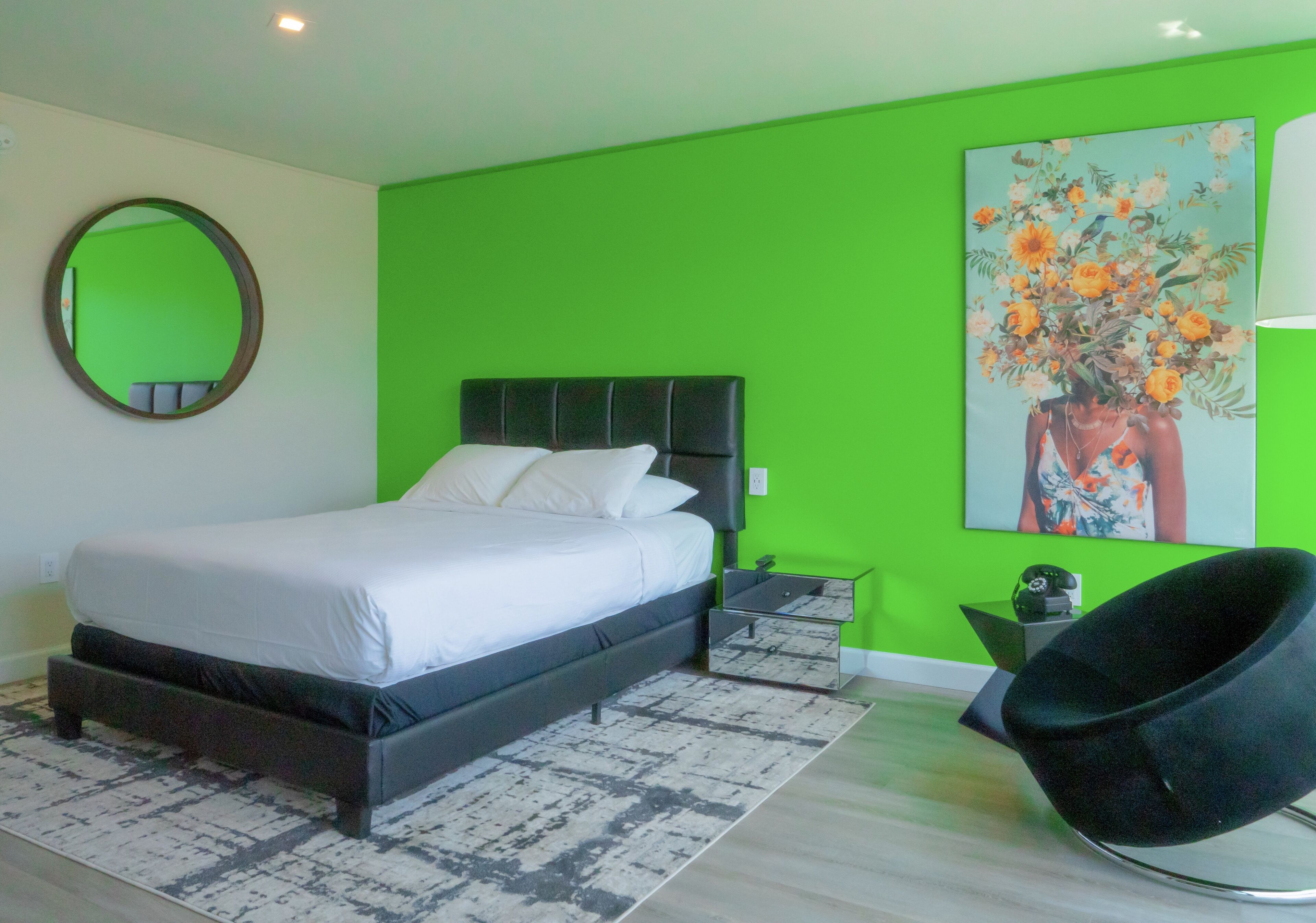 Quarto moderno com parede verde de destaque, cama com roupa de cama branca, cabeceira preta, espelho redondo, mesa de cabeceira, cadeira preta e uma obra de arte floral.