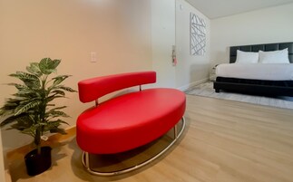Une chambre moderne avec un fauteuil rouge, un lit recouvert de linge de lit blanc, une plante en pot et une œuvre d’art géométrique accrochée au mur.