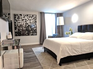 Een moderne hotelkamer met een groot bed, een glazen tafel, een stoel, een bureau, een groot abstract schilderij en een raam met gordijnen.