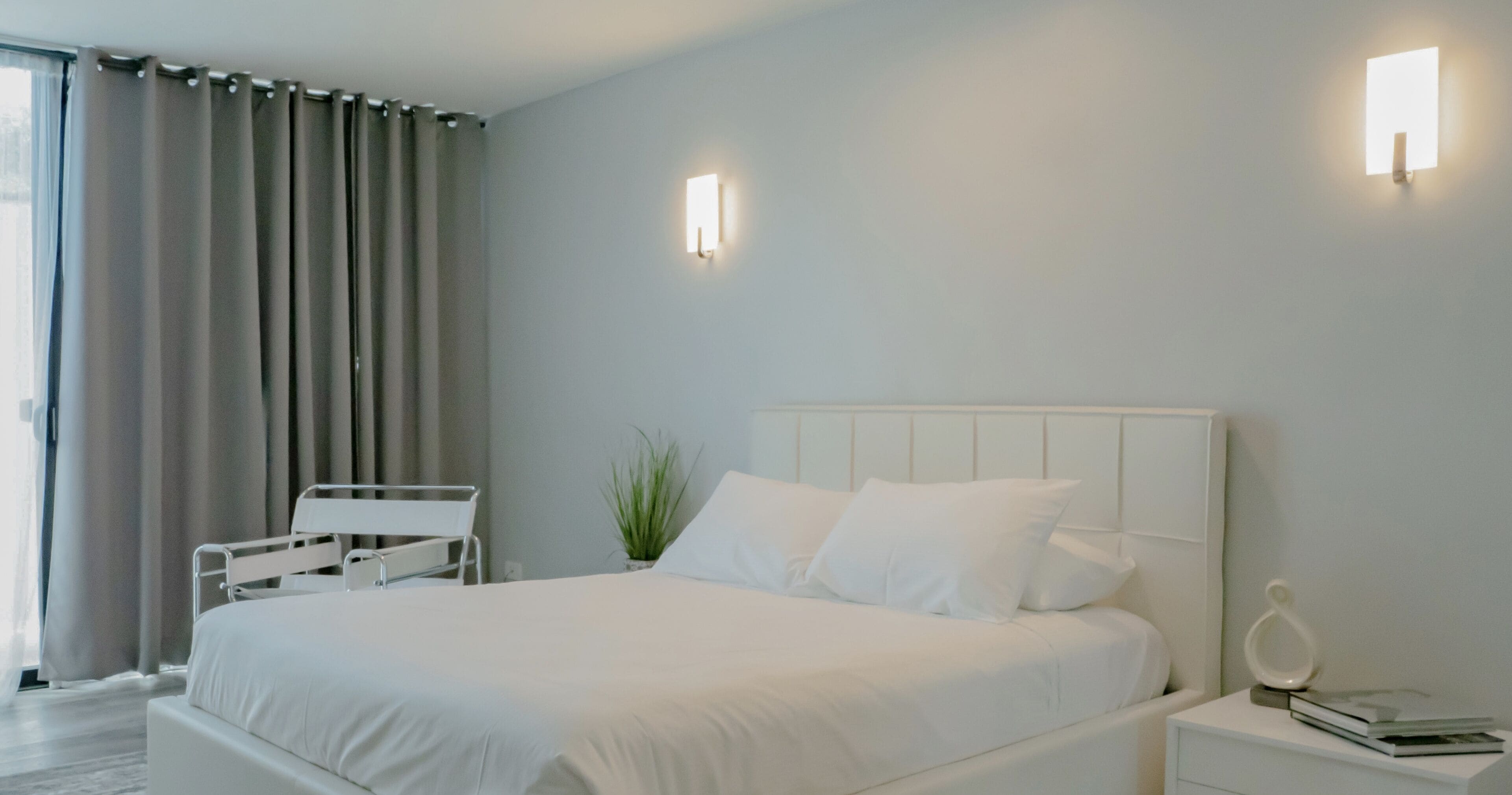 Un dormitorio moderno con una cama grande, mesitas de noche, una mesita auxiliar, una planta en maceta y una ventana con cortinas.