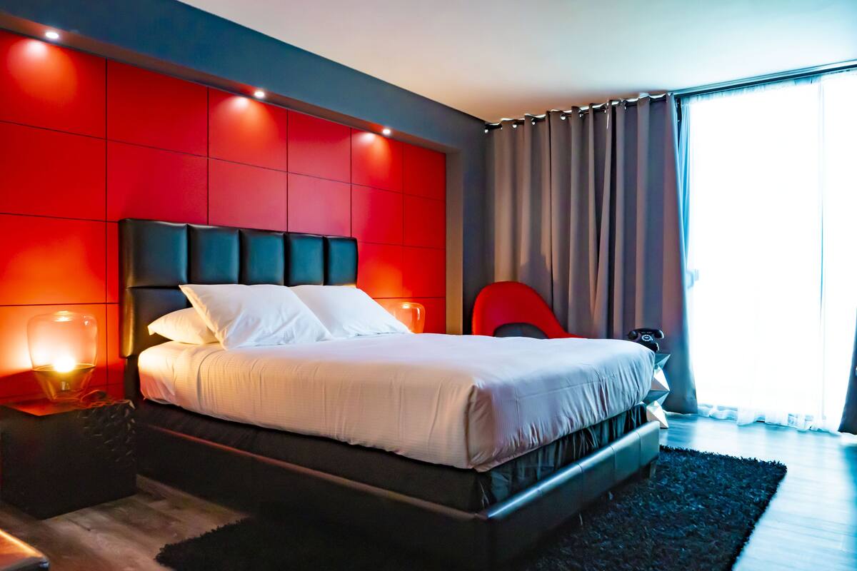 A modern bedroom with a large bed, red accent walls, and a view of the outdoors through a window.