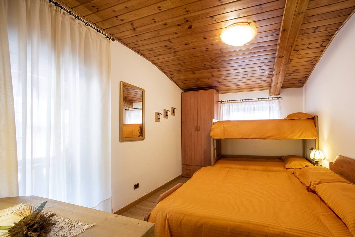 A room with wooden ceiling beams, a bunk bed with orange bedding, a wooden wardrobe, a mirror, and a small table with a decorative item.
