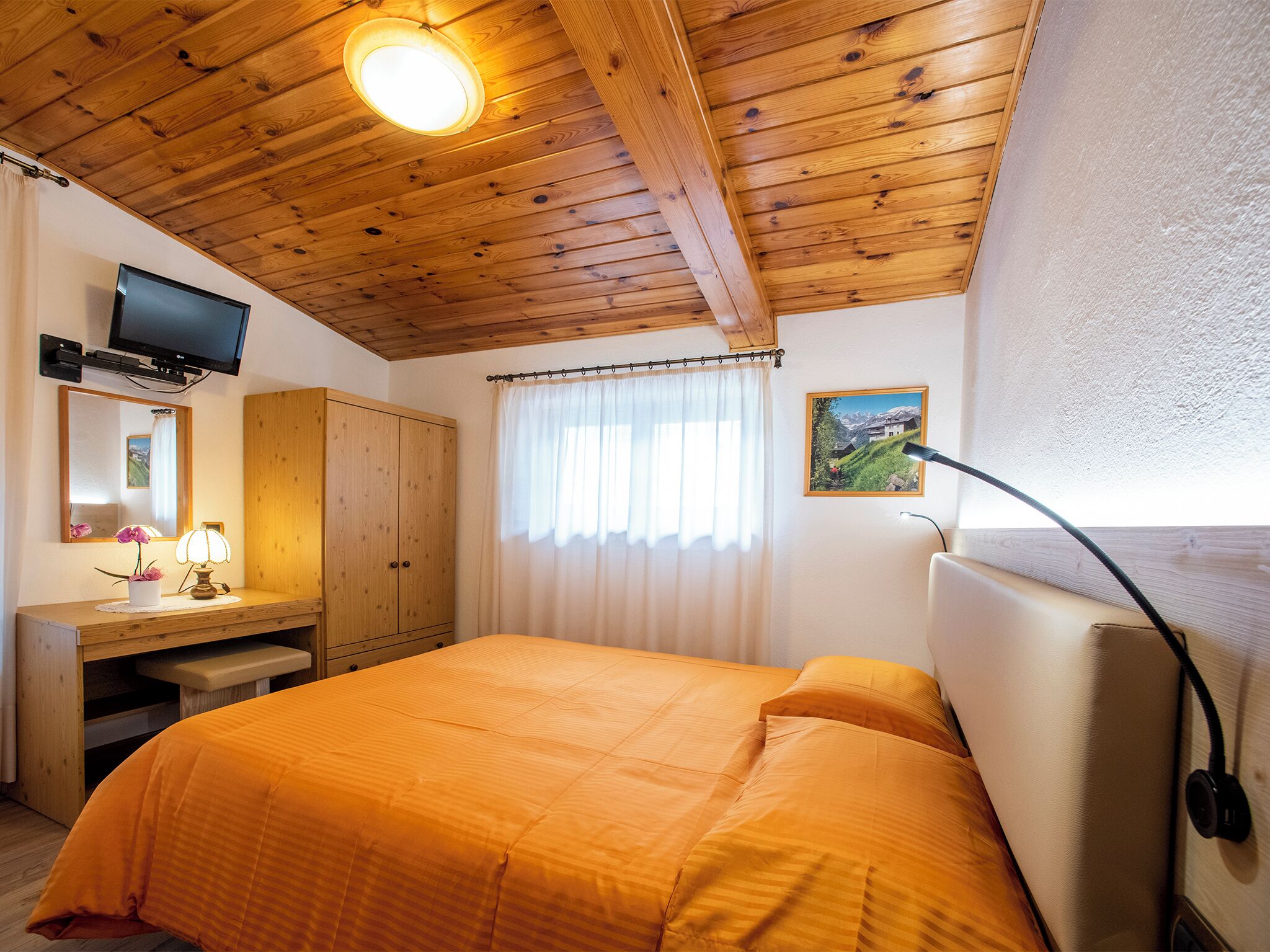 Ein Schlafzimmer mit Holzboden, einem Bett mit orangefarbener Bettdecke, einem Schreibtisch mit Lampe, einem Fernseher und einem Bild an der Wand.