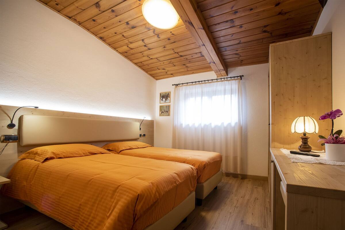 A bedroom with two beds, wooden ceiling, a nightstand, a lamp, and a small desk with a flower.